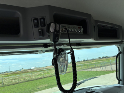 Truck cab interior showing CB radio mic and control panel mounted over windshield with vents and switches in view; exterior field visible through front window.