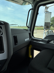 Truck cab interior showing dashboard with a small label near the glove box and a yellow handle, window and side mirror, view of outdoors.