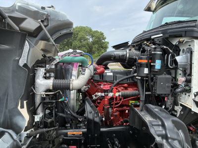 Truck engine bay showing Mack GR64F diesel engine with red components and various hoses; visible air intake, cooling system, and wiring harnesses.