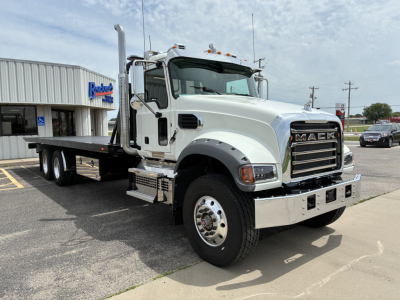 Mack GR64F tow truck in a dealership lot showing front grille with Mack logo, chrome bumper, dual exhaust stacks, white cab, flatbed rollback body, and side steps.
