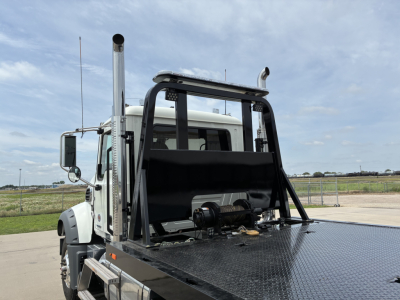 Truck cab with a metal flatbed and roll-back winch apparatus, tall exhaust stacks, and protective rollover bars. Light-colored truck in open lot.