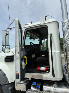 Truck cab interior visible through open door of a white Mack GR64F sleeper. Steering wheel, driver's seat, and control panels visible; blue bottle and red fire extinguisher in the step area.