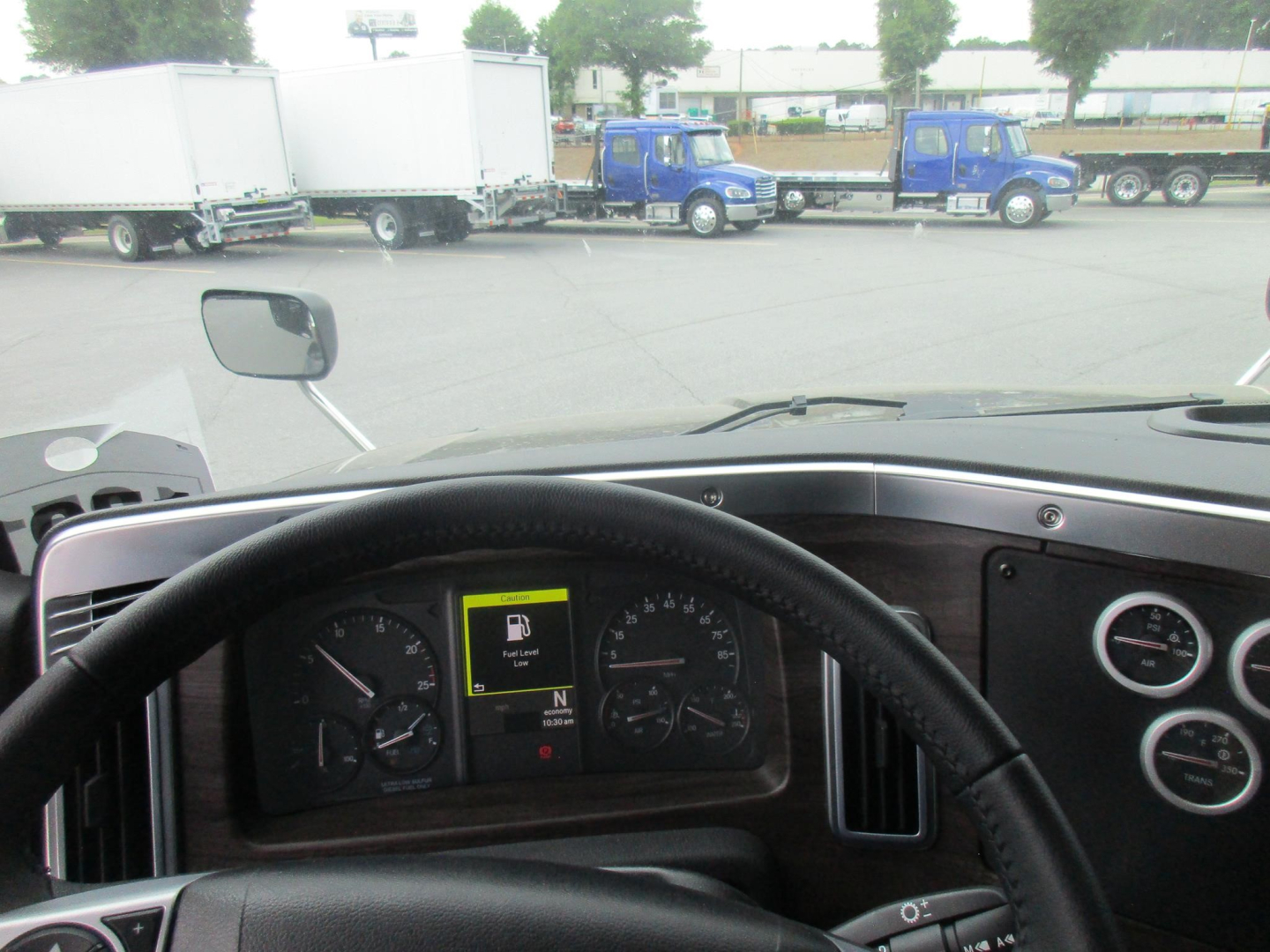 Cab interior of a Western Star truck showing steering wheel, dashboard, and instrument cluster with center display; exterior lot with blue trucks and trailers in background.