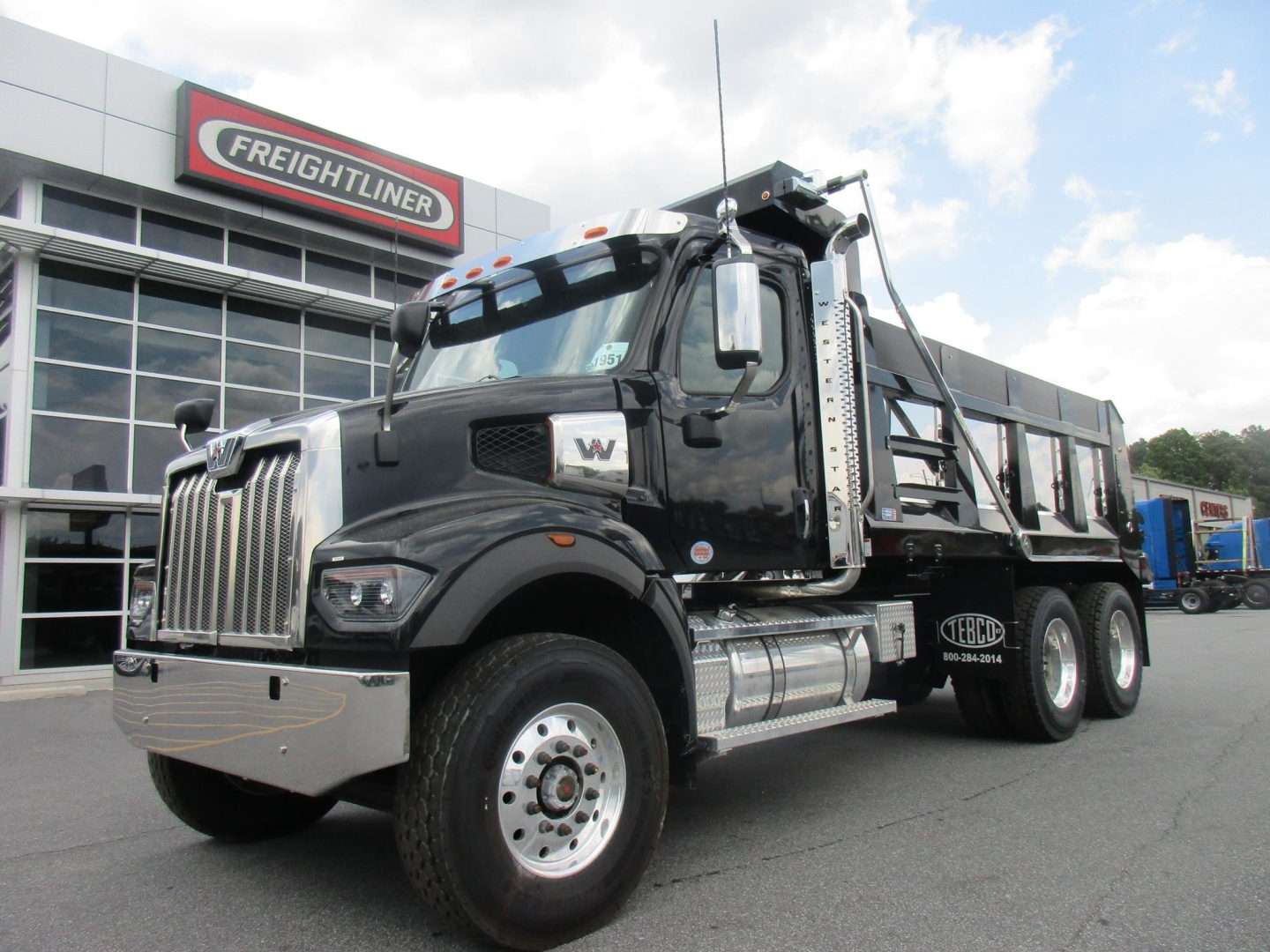Western Star 47X dump truck in front of Freightliner dealership showing cab, chrome bumper, dual rear wheels, and dump body with ladder rack.