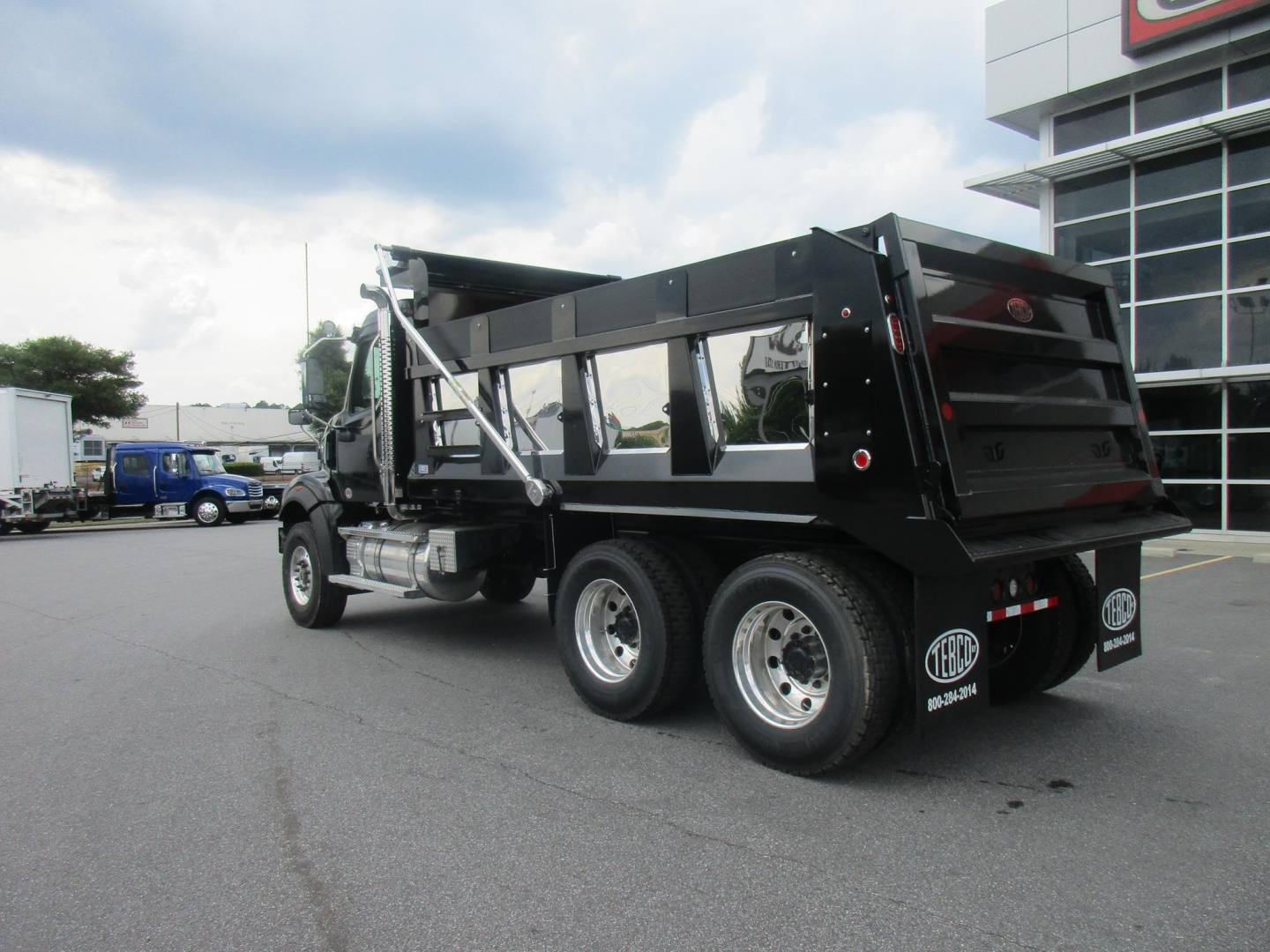 Truck: 2027 Western Star 47X dump truck with black dump body, dual rear wheels, chrome fuel tanks, and side steps; buildings and other trucks in background.
