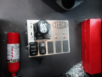Small control panel labeled TEBCO with toggle switches and a key, two warning lights, a fire extinguisher on left, and a red safety box on right. Visible text includes TEBCO and various switch labels.