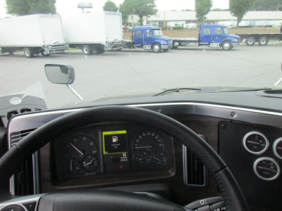 Cab interior of a Western Star truck showing steering wheel, dashboard, and instrument cluster with center display; exterior lot with blue trucks and trailers in background.
