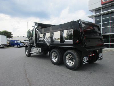 Truck: 2027 Western Star 47X dump truck with black dump body, dual rear wheels, chrome fuel tanks, and side steps; buildings and other trucks in background.