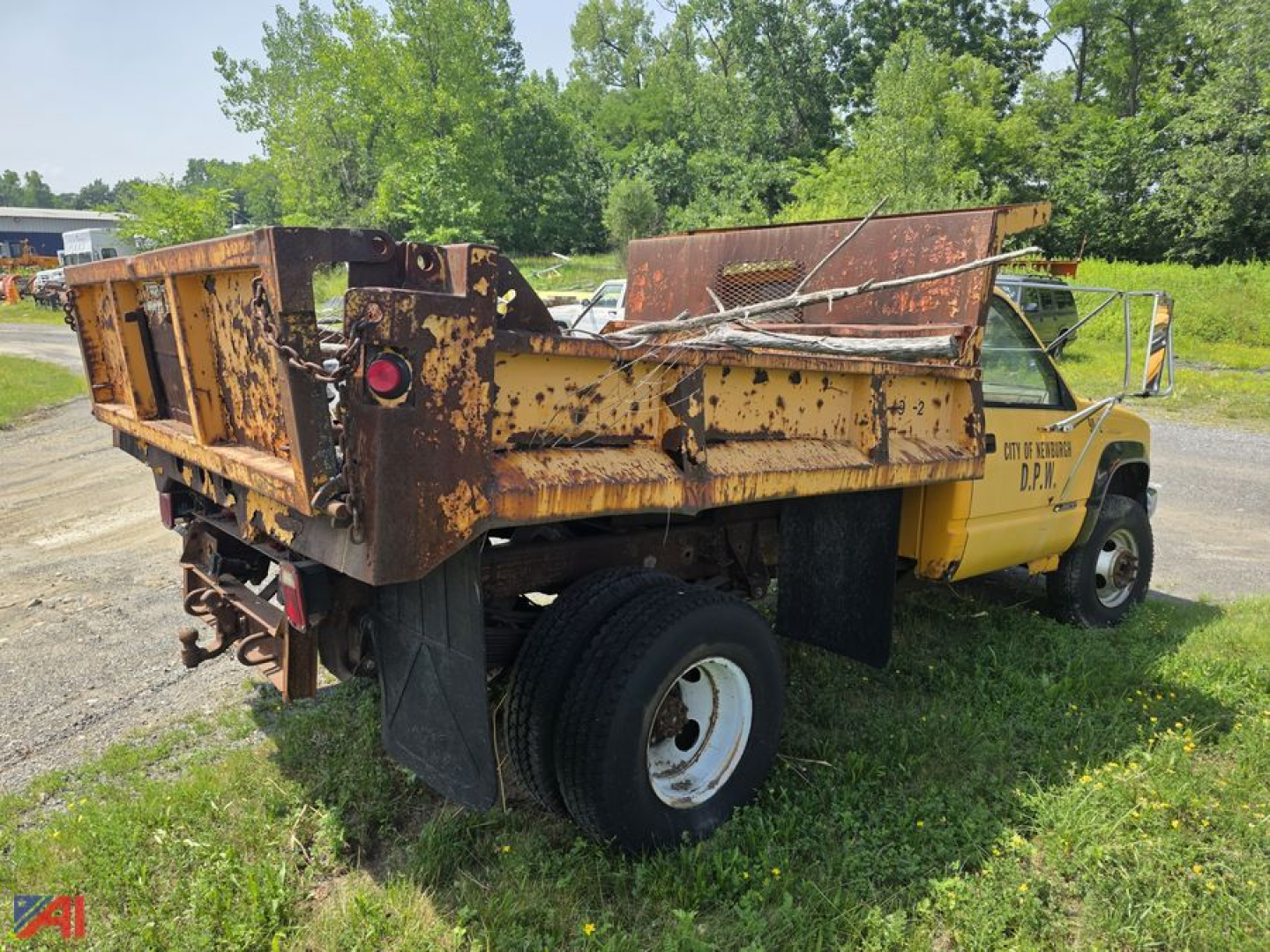 Dump truck rear view showing rusted steel dump bed and yellow cab with D.O.T. markings, single rear axle, visible hydraulic components, and in-service tires.