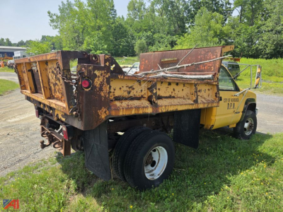 Dump truck rear view showing rusted steel dump bed and yellow cab with D.O.T. markings, single rear axle, visible hydraulic components, and in-service tires.