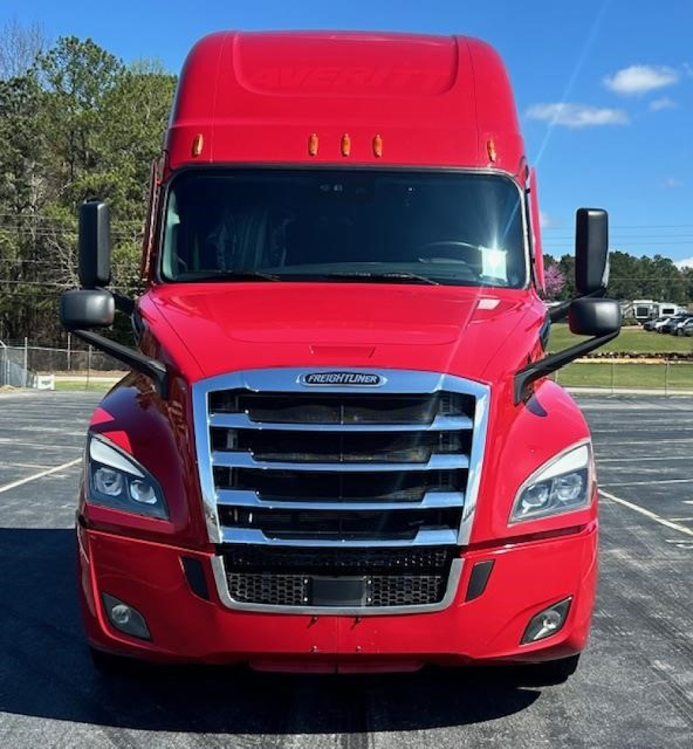 Front view of a red 2023 Freightliner Cascadia 126 tractor-trailer in a parking lot showing grille, headlights, and bumper.