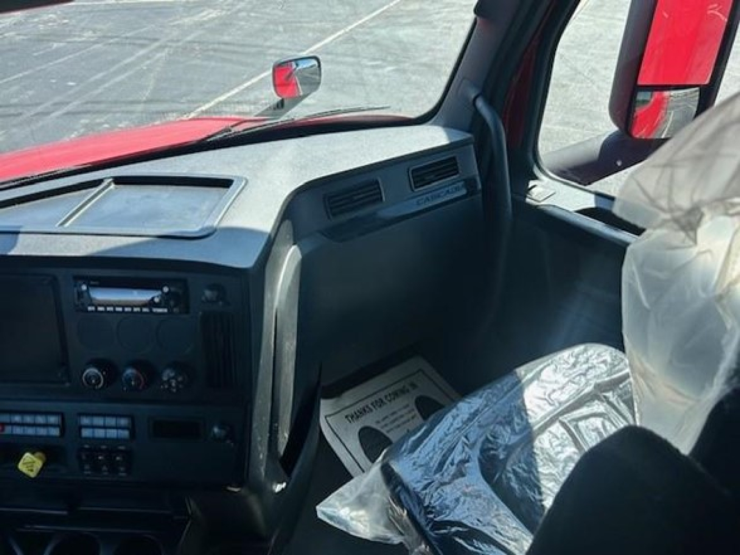 Truck cab interior showing dashboard controls and steering wheel area of a Freightliner Cascadia. Visible are HVAC dials, radio, and storage panel, with plastic-wrapped seats.