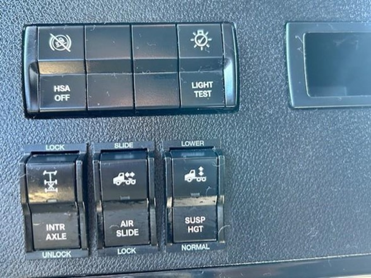 Dashboard switches and controls of a Freightliner Cascadia showing HSA OFF, LIGHT TEST, LOCK, SLIDE, LOWER, INTR AXLE, AIR SLIDE, SUSP HGT, NORMAL.