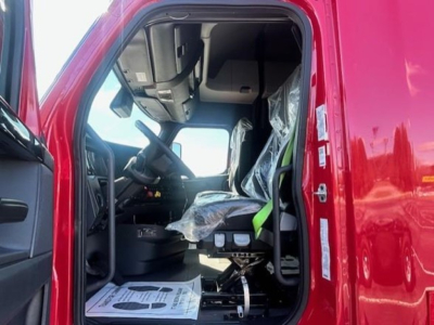 Truck cab interior of a Freightliner Cascadia 126 from driver’s side showing steering wheel, seat, and dashboard with plastic-covered seats.
