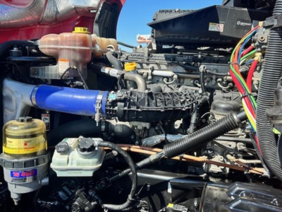 Freightliner Cascadia 126 engine bay showing coolant reservoir, blue intercooler hose, hydraulic fluid tank, wiring harness, and diesel engine components.