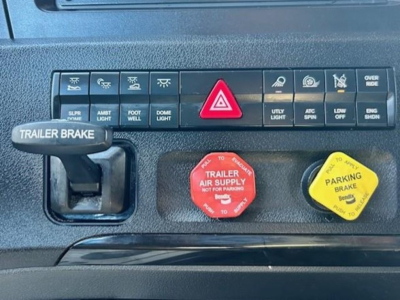 Dashboard controls of a Freightliner Cascadia showing trailer brake lever, red emergency/air supply knob, yellow parking brake knob, and labeled function switches.