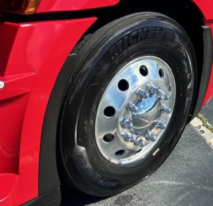 Front wheel of a red Freightliner Cascadia sleeper showing Michelin tire and polished multi-hole chrome hub with lug nuts.