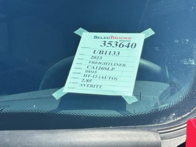 Truck window showing a green tag with listing details: STOCK 353640, UB1133, 2023 Freightliner Cascadia, CA126SLP, DD15, DT-12 (AUTO), 2.85, VERITIT