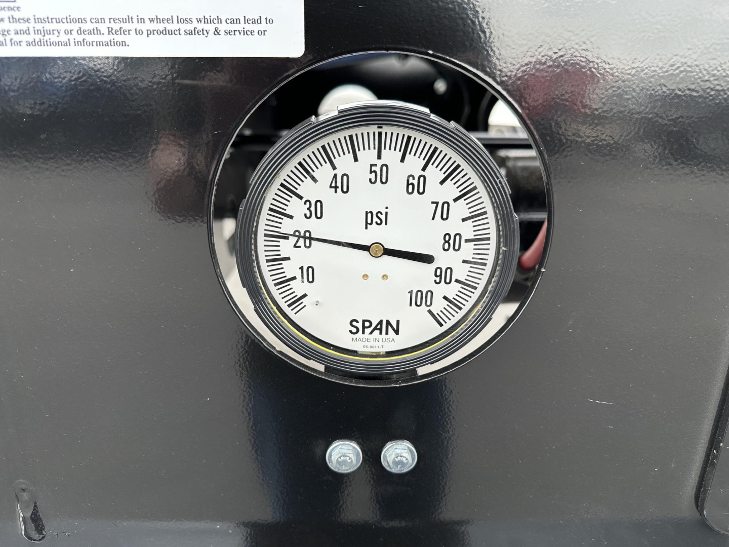 Gauge showing pressure in psi from 0 to 100 on a circular dial with 'SPAN' marking at bottom, mounted on a dark trailer surface with two small bolts below.