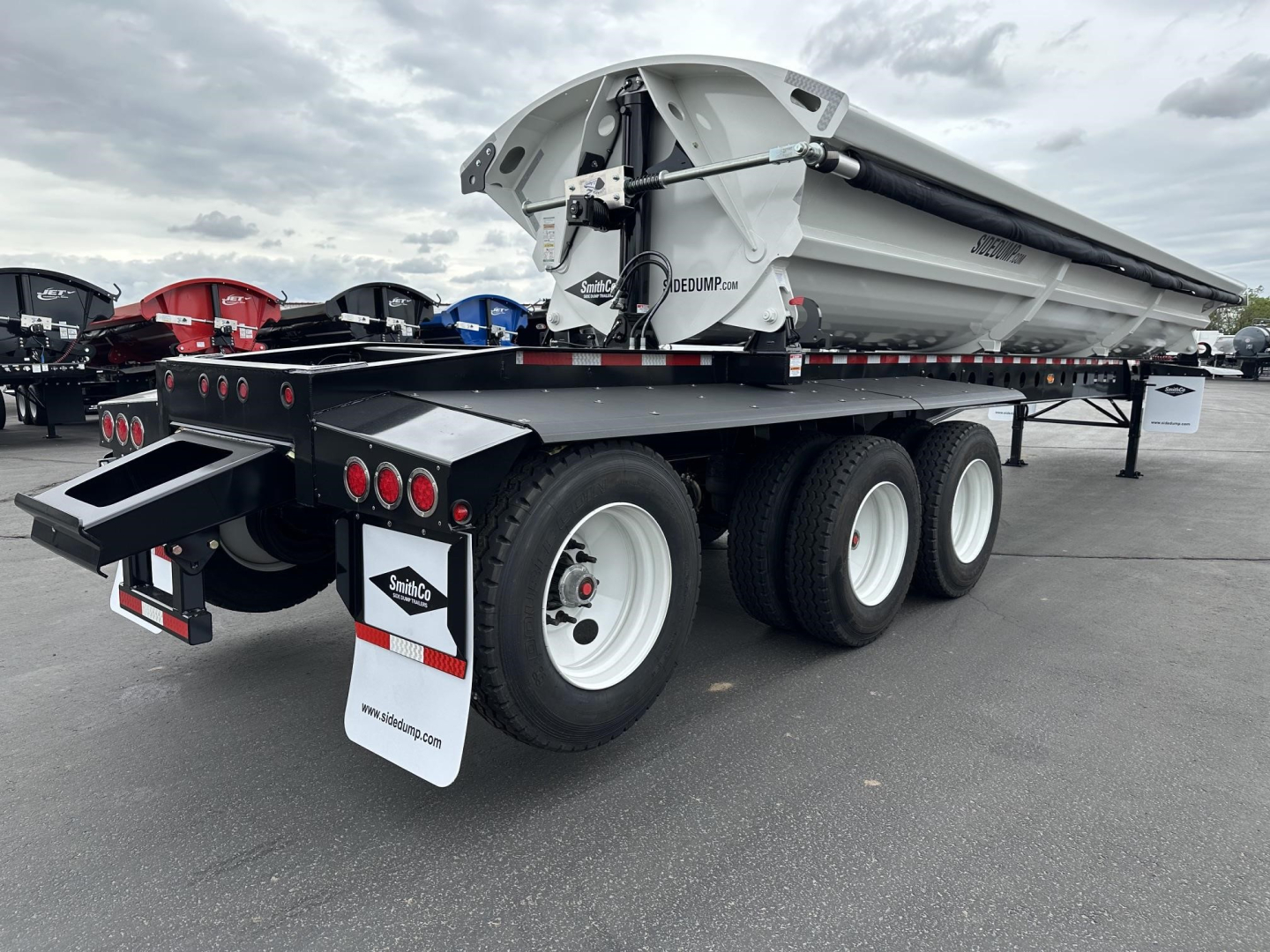 Trailer rear view of a Smithco Side Dump W Super Spread trailer with dual rear axles, white wheels, black chassis, and a folded dump bed. Visible text includes Smithco branding on mudflaps and hopper.
