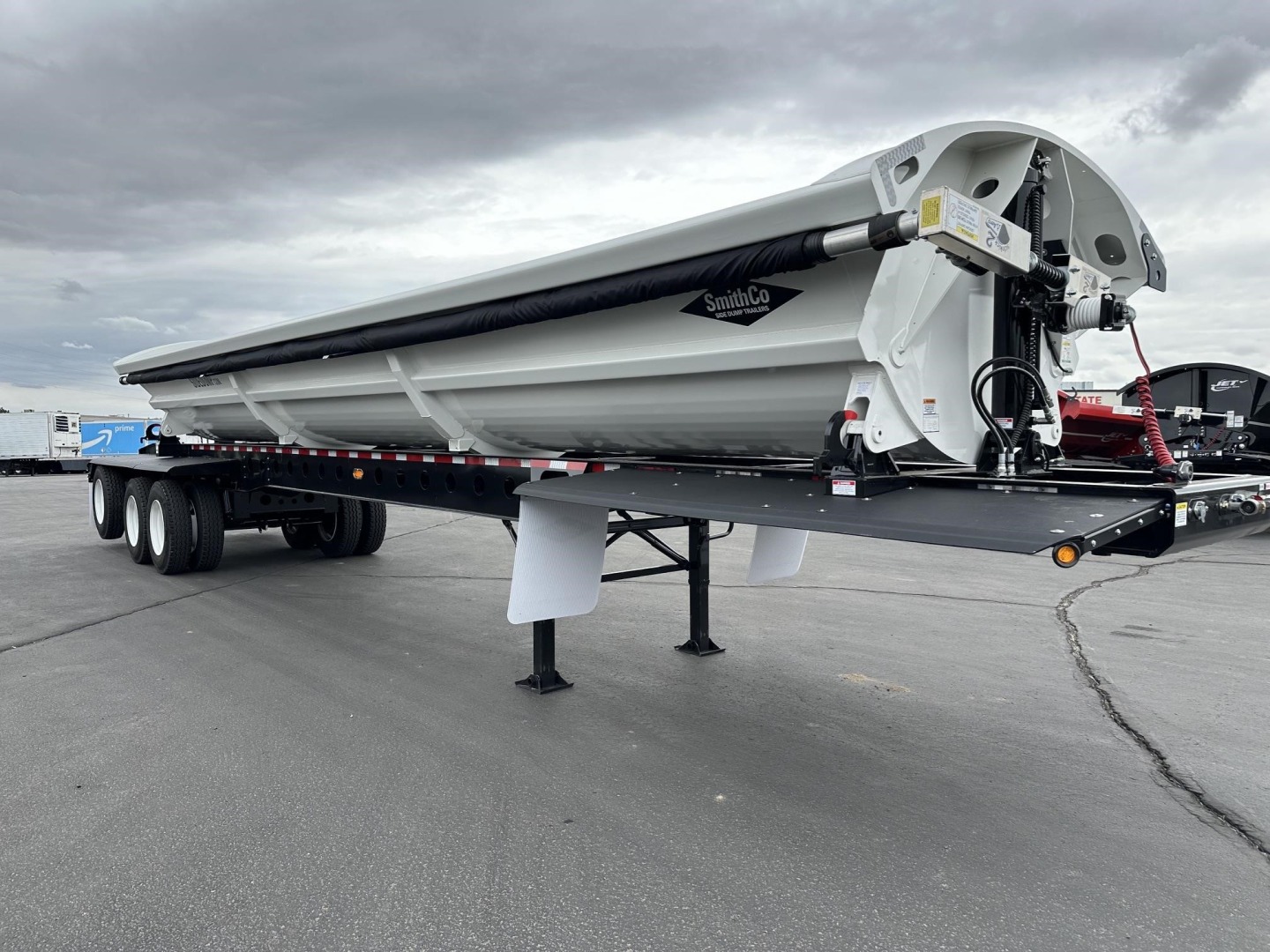 Side dump trailer in profile showing folded auger and hydraulic valve, SmithCo side dump logo, white and black color scheme, hitch area and dual rear axles.