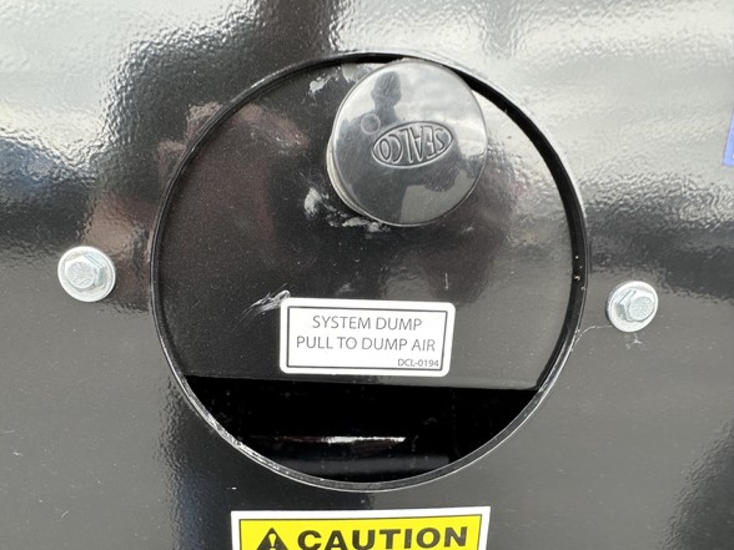 Side view of a new Smithco tri-axle dump trailer showing a round access hatch with a locking cap and a CAUTION label near the trough.