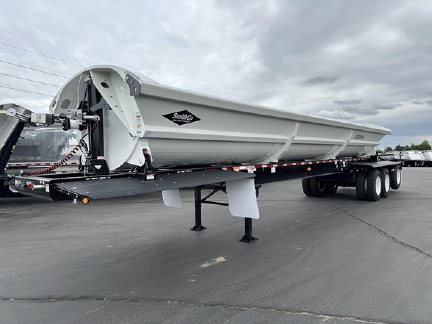 Smithco tri-axle side dump trailer with galvanized gray body, front hitch, hydraulic cantilever hoist, and dual rear axles on a black asphalt lot.