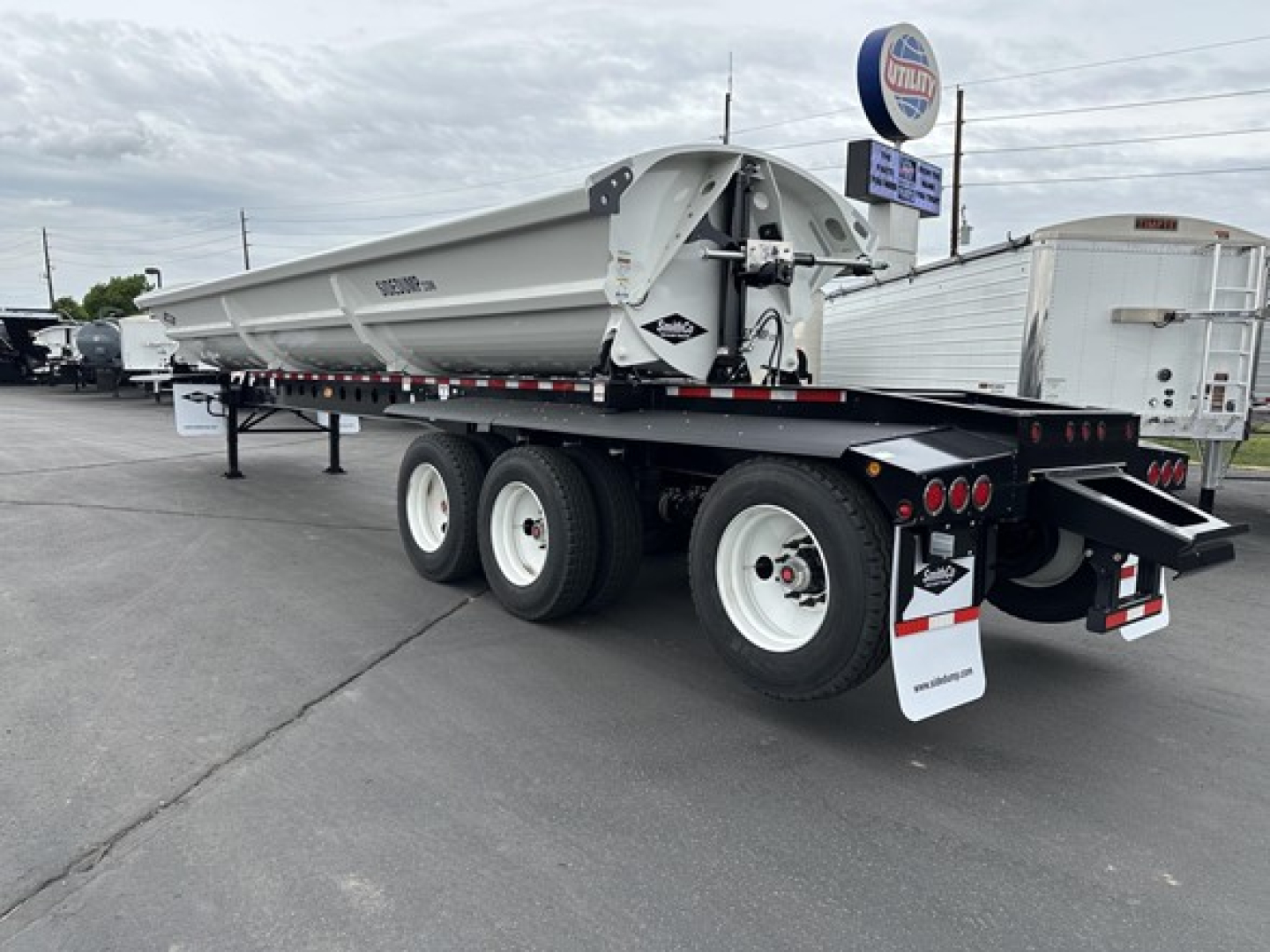 Tri-axle side dump trailer visible in a lot, tandem axle setup with three axles, gray dump body, rear tailgate, black frame, and white wheels.
