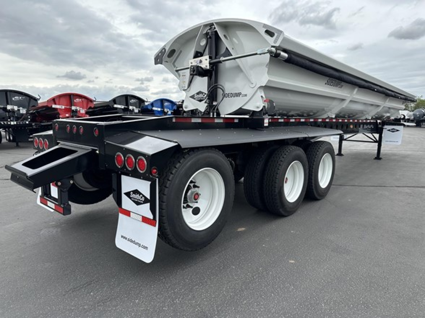 Tri-axle side dump trailer showing a large metal dump bed and multiple axles with white wheels; rear lights, mud flaps, and chassis visible; SmithCo branding on bed.