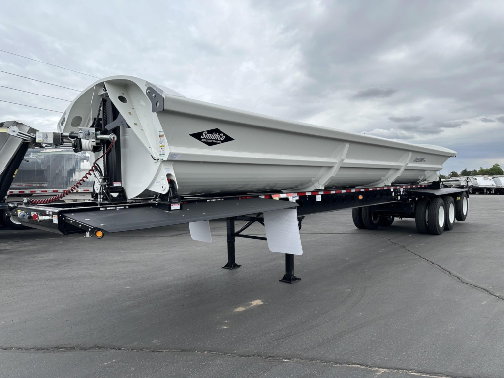 Smithco side dump trailer with spreader mechanism visible, side railing, white/gray body, logo reads Smithco on side panel. Trailer sits on triple-axle configuration with landing legs in front.