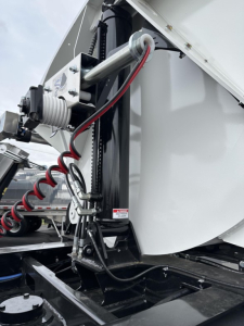 Trailer hitch area showing hydraulic cylinder and coiled air hose attached to a silver and black dump trailer body. Visible text on a small label near the hydraulic component reads 'A...'.