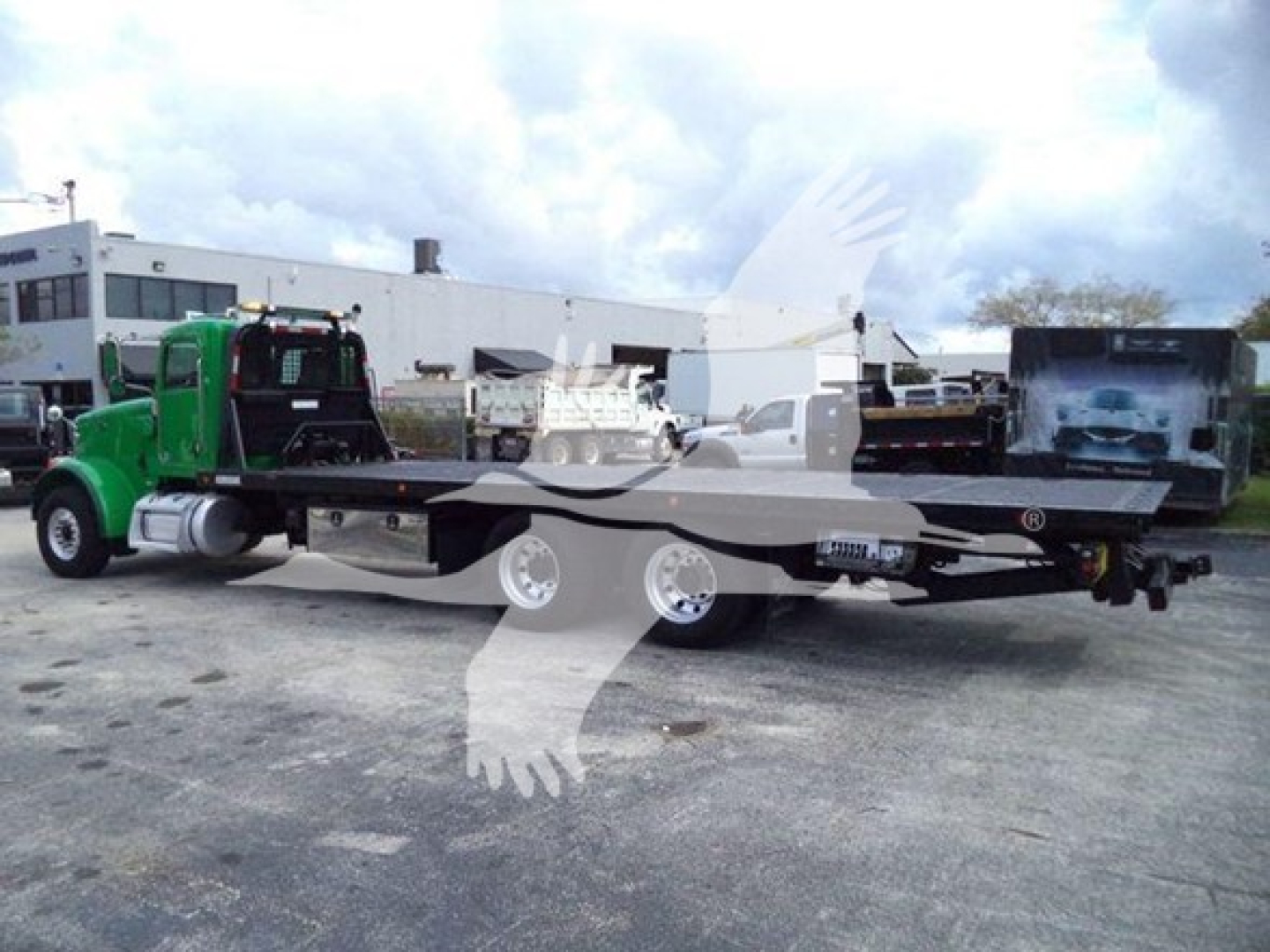 Flatbed rollback tow truck with a green cab and long flatbed, viewed from the side in an outdoor lot.