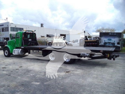 Flatbed rollback tow truck with a green cab and long flatbed, viewed from the side in an outdoor lot.