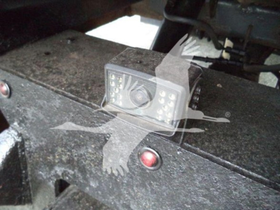 Truck camera or sensor mounted on chassis showing a small rectangular device with multiple LEDs and a central lens; surface around is textured metal with dirt.