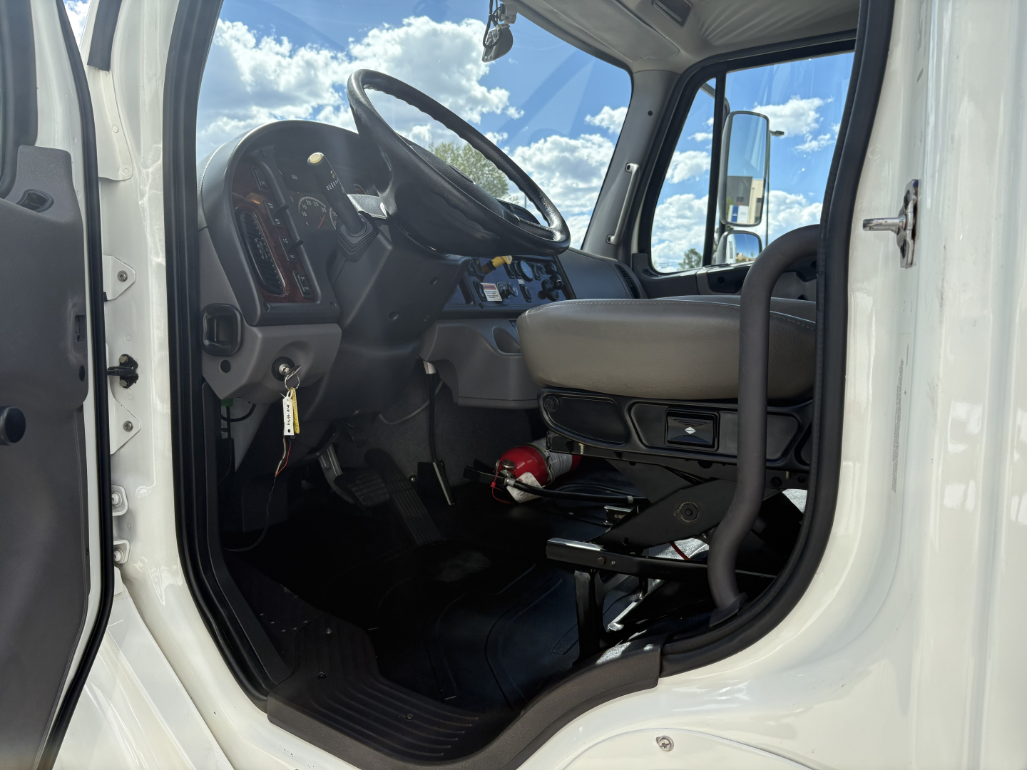 Interior of a 2022 Freightliner M2-106 cab showing steering wheel, dashboard, and driver seat with a red fire extinguisher mounted under the dash.