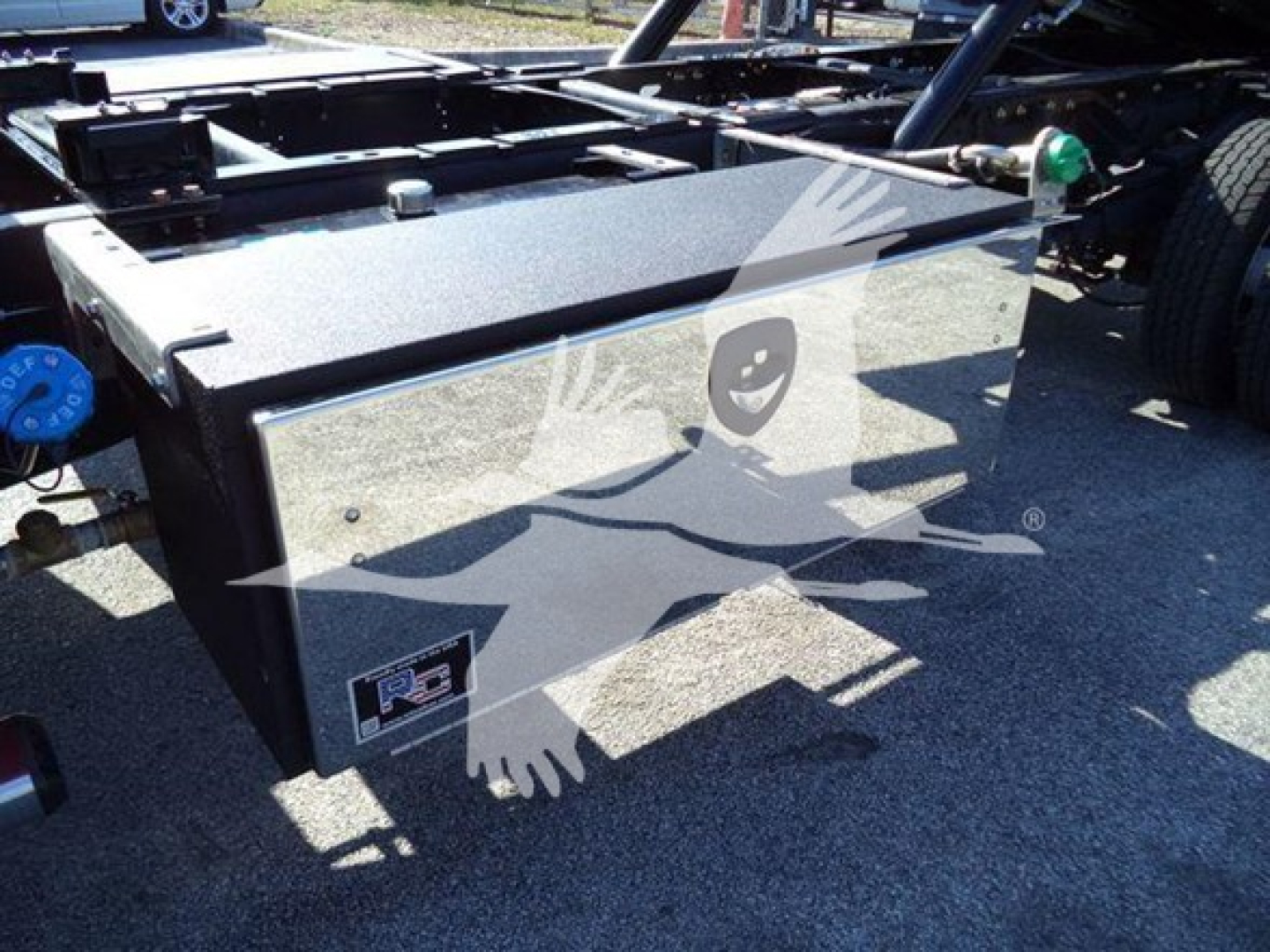 Truck chassis with fuel tank enclosure and dual rear wheels visible; side view shows large rectangular fuel tank/box with metal surface and a small label at lower left; blue cap on left and green marker light on right edge.