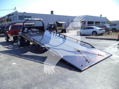 Ram 5500 rollback tow truck flatbed with ramp lowered, rear view showing long steel deck and wheel chock, dealership yard in background.
