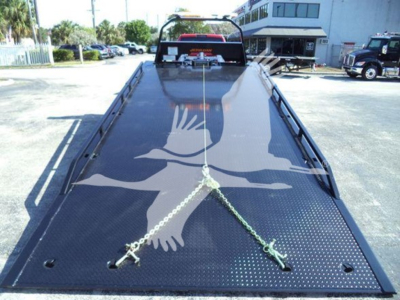 Ram 5500 rollback flatbed truck bed with black perforated steel decking and chains securing a vehicle, rear view with cab and yard in background.
