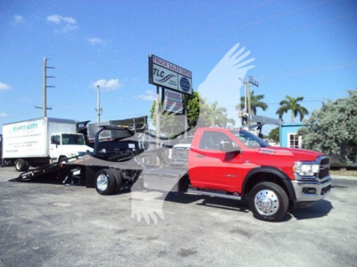 Red Ram 5500 rollback tow truck with a black tilt bed trailer in a dealership lot. Signage in background reads 7LC, with palm trees and a blue sky.