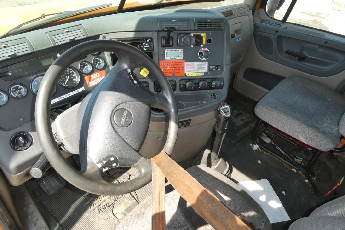Truck cab interior showing steering wheel, dashboard with gauges, gear shifter, and driver seat. Visible labels and switches on center console, some warning stickers.
