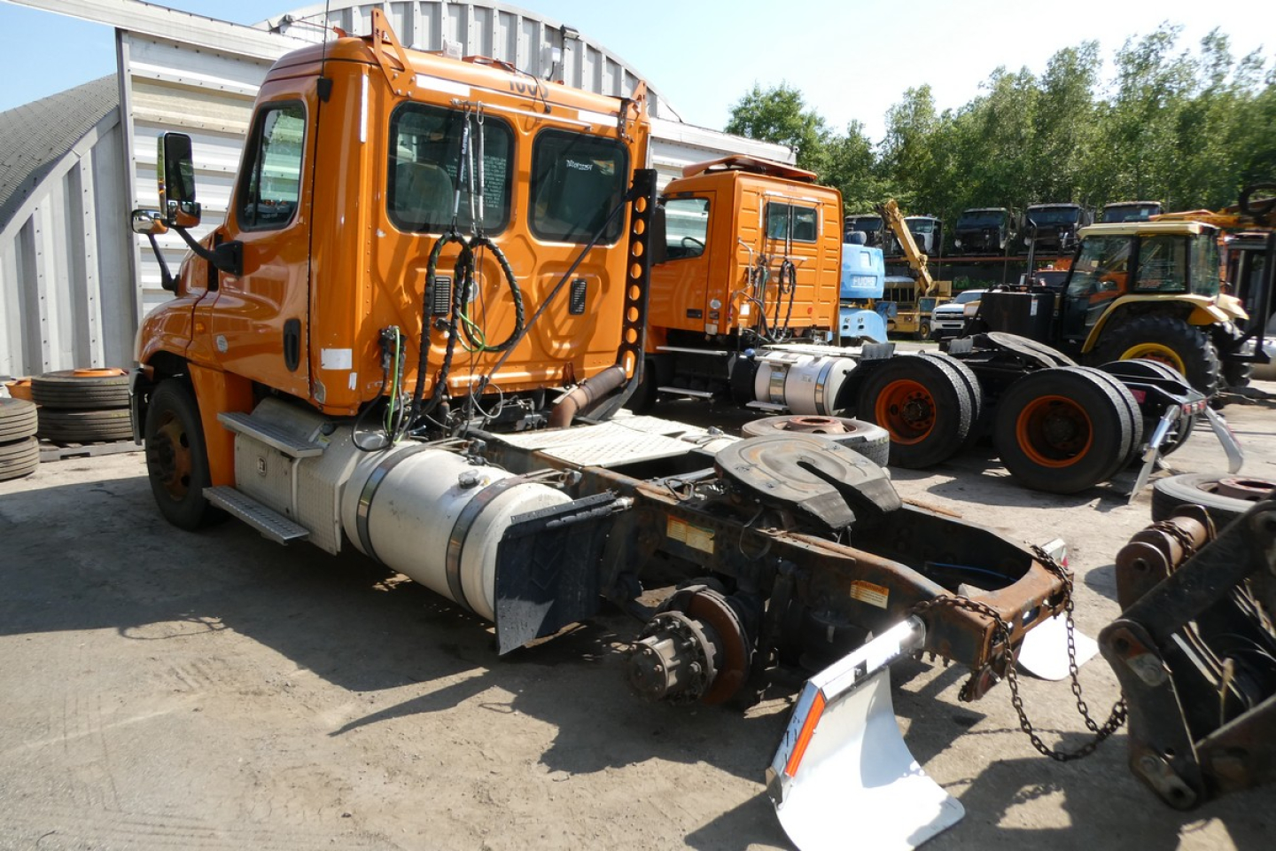 Orange Freightliner Cascadia 125 daycab truck with missing rear driveshaft area, side view shows cab and chassis in a yard with other trucks.