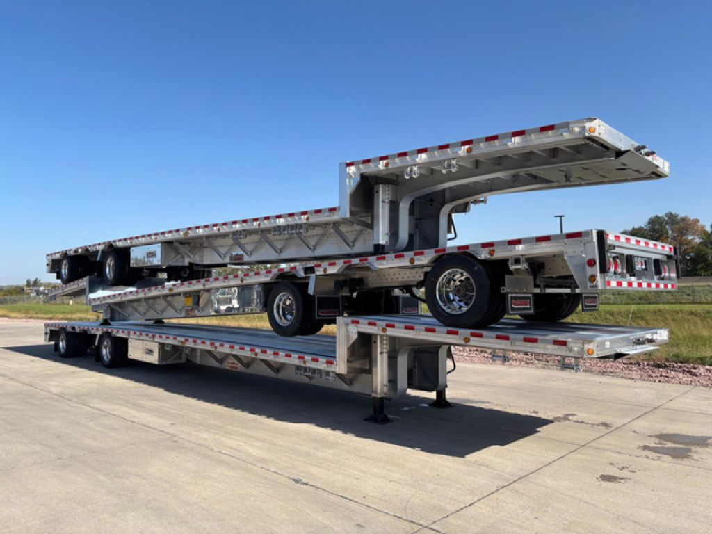 Dropdeck trailer with stacked sections and multiple axles, deck rails, and reflective safety striping; aluminum frame, ramps folded on top; side view.
