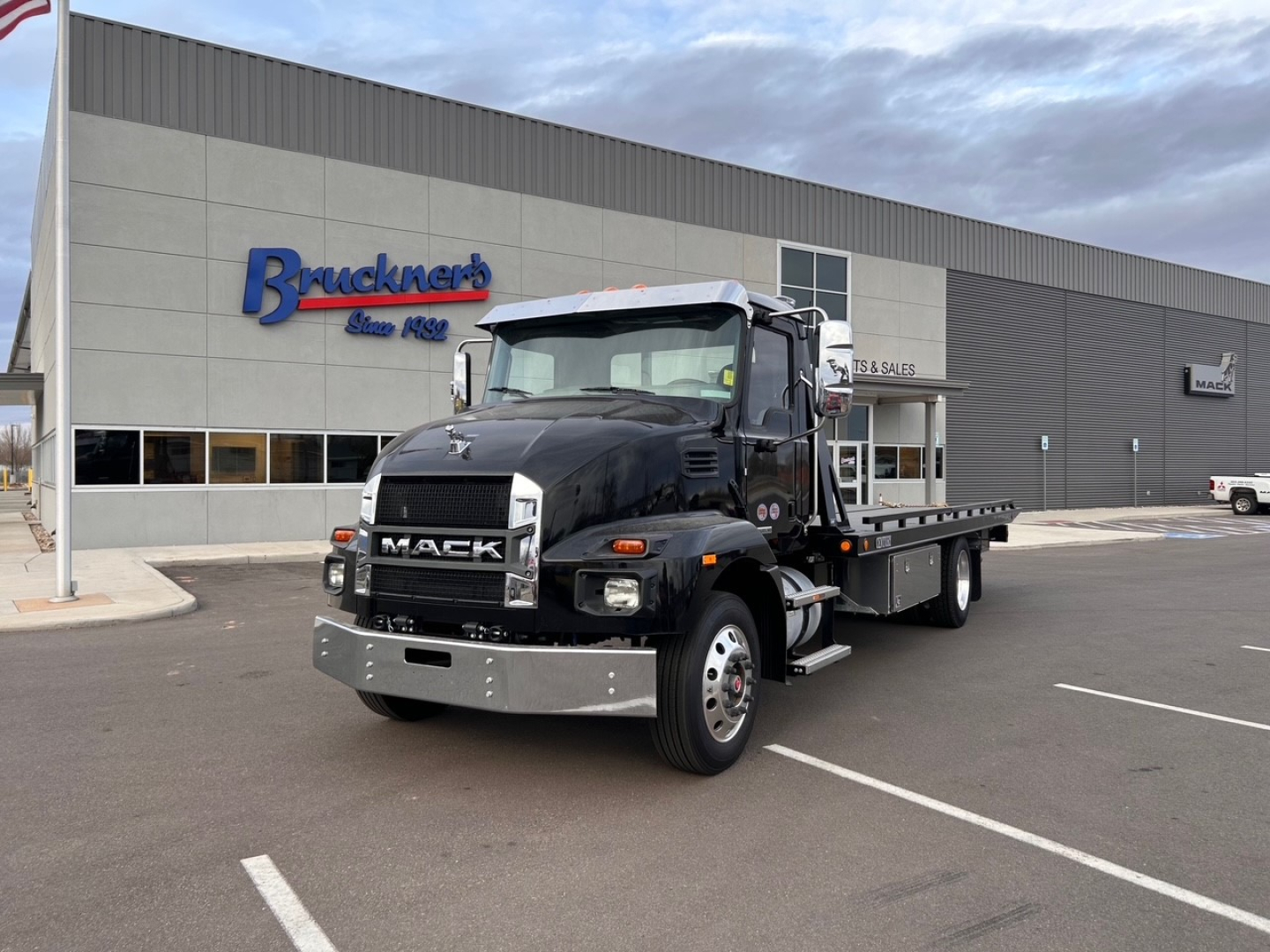 Black 2025 Mack MD6 tow truck in front of a dealership, bold grille with MACK logo, flatbed rear, daytime scene, clear sky.
