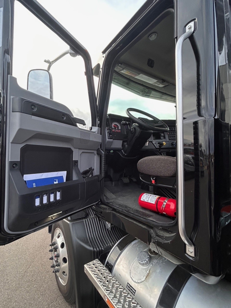 Truck cab interior and entry from driver side showing open door, dashboard, steering wheel, and fire extinguisher in footwell.