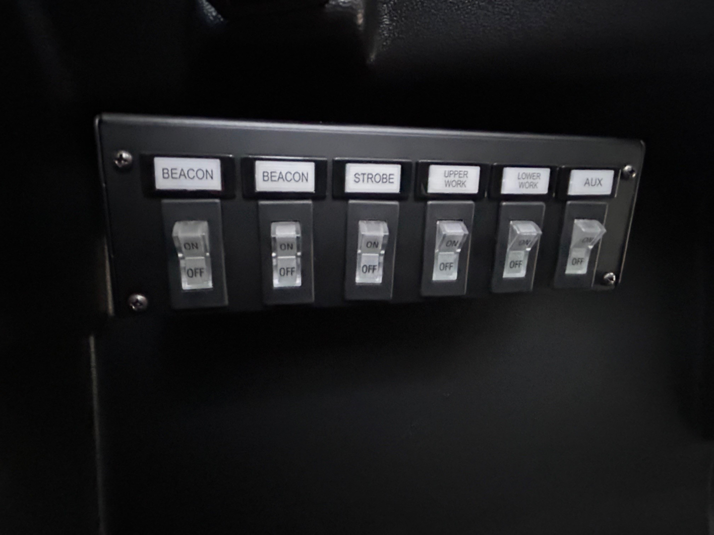 Panel of switches labeled BEACON, BEACON, STROBE, UPPER WORK, LOWER WORK, AUX in a Mack MD6 truck cab showing control switches.