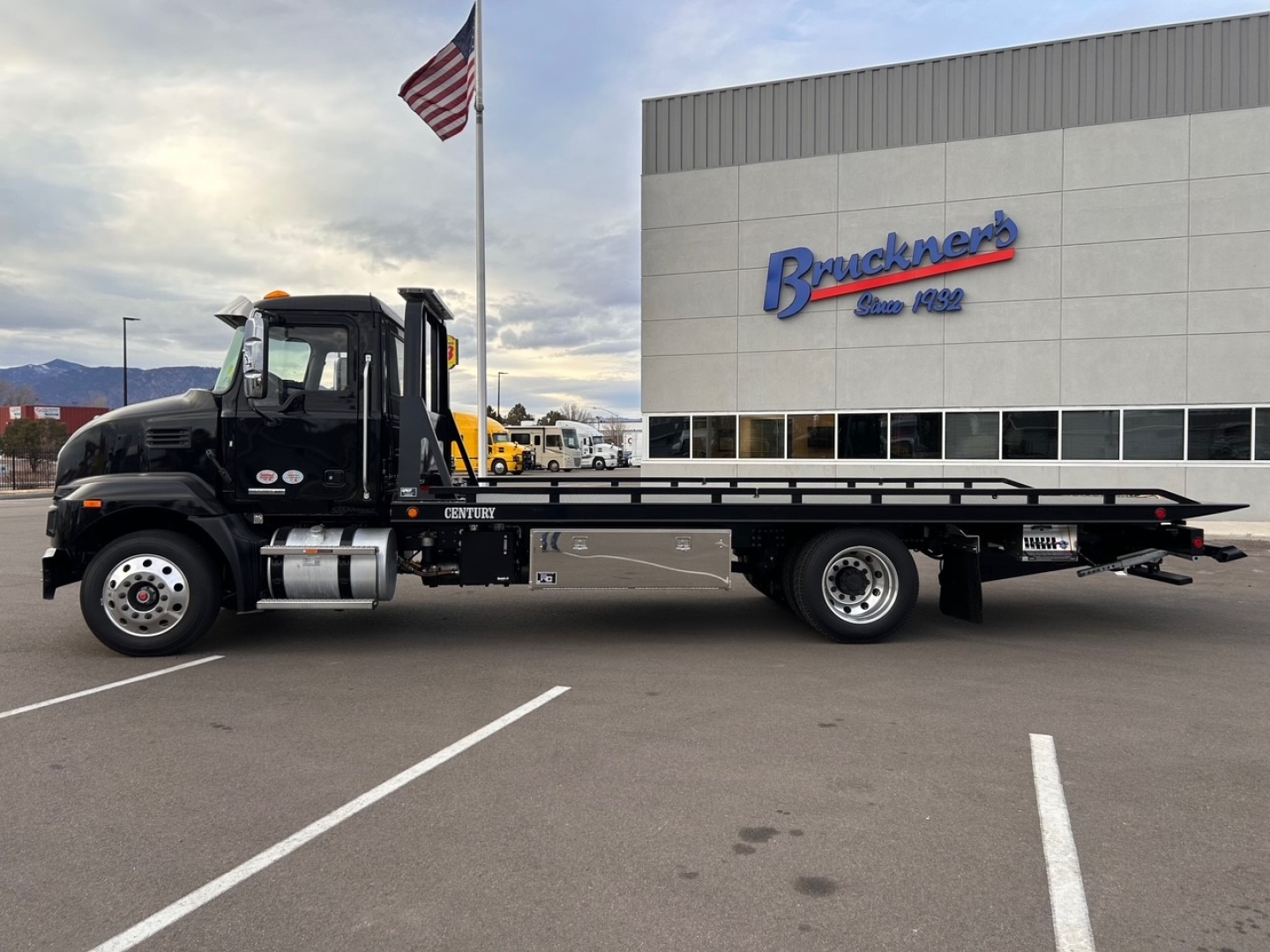 Mack MD6 tow truck in profile view showing flatbed and cab with Bruckner sign building in background.