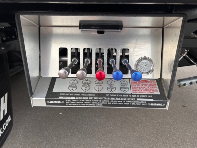 Truck control panel with six levers labeled and a dial gauge on the right. See red, blue, and silver levers and warning labels below the levers.