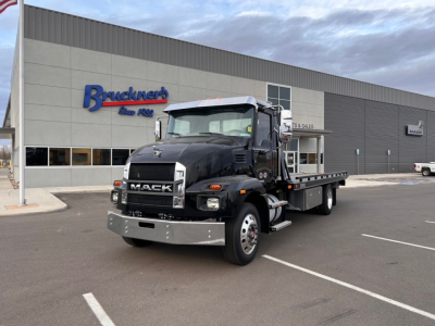 Black 2025 Mack MD6 tow truck in front of a dealership, bold grille with MACK logo, flatbed rear, daytime scene, clear sky.