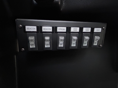 Panel of switches labeled BEACON, BEACON, STROBE, UPPER WORK, LOWER WORK, AUX in a Mack MD6 truck cab showing control switches.