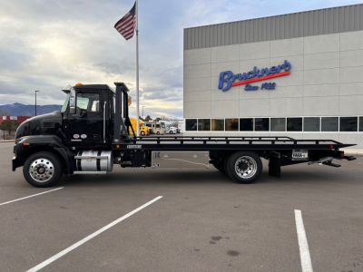 Mack MD6 tow truck in profile view showing flatbed and cab with Bruckner sign building in background.